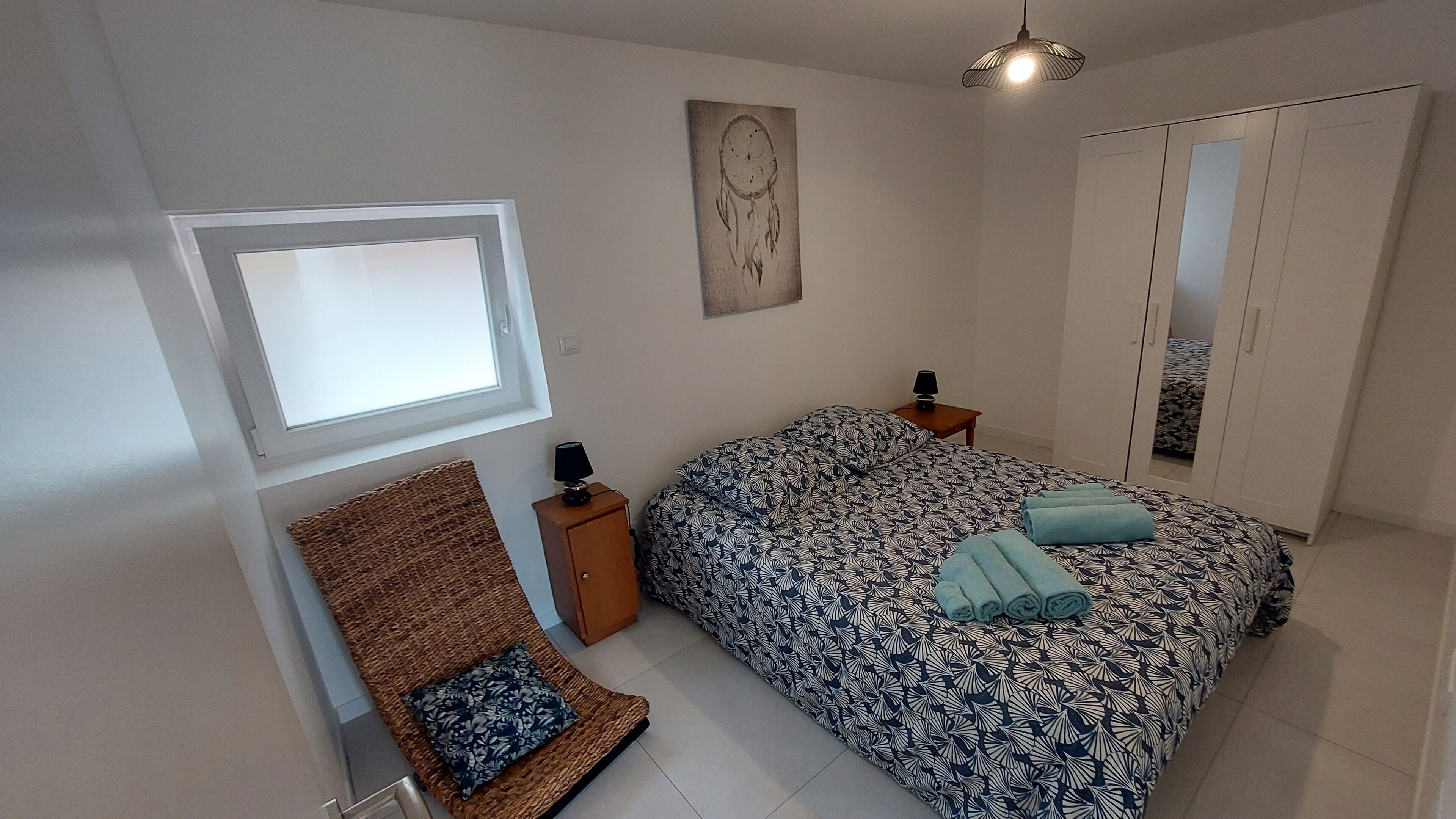 A bedroom with a bed, a nightstand, a lamp, a woven chair, a wall art piece, and a wardrobe.