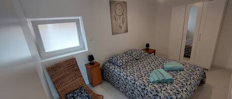 A bedroom with a bed, a nightstand, a lamp, a woven chair, a wall art piece, and a wardrobe.
