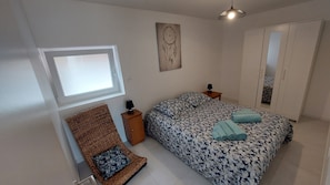 Une chambre à coucher comprenant un lit, une table de chevet, une lampe, une chaise en osier, une œuvre d’art murale et une armoire.