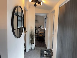 Un couloir menant à une cuisine, où l’on trouve un miroir, une plante en pot et un paillasson.