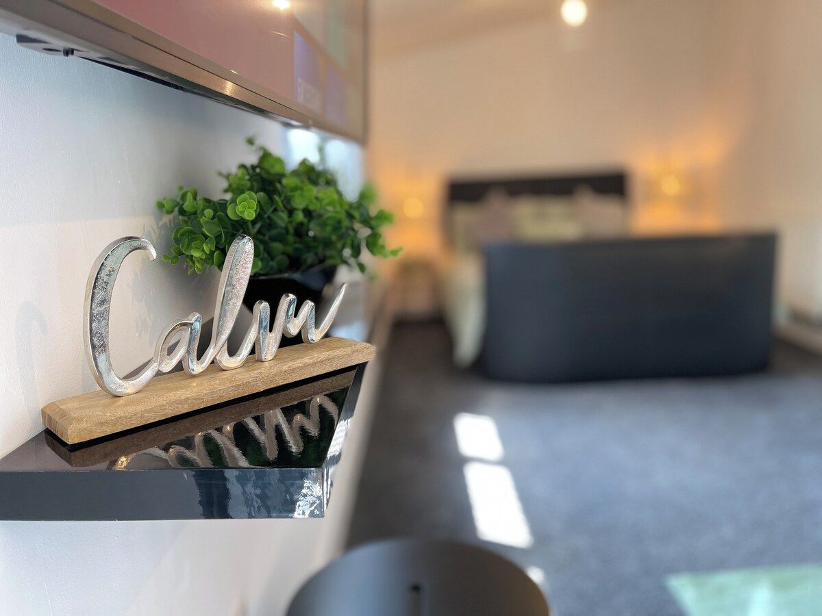 Ein modernes Interieur mit einem dekorativen Schild, auf dem 'Calm' steht, einer Topfpflanze und einer Couch im Hintergrund.