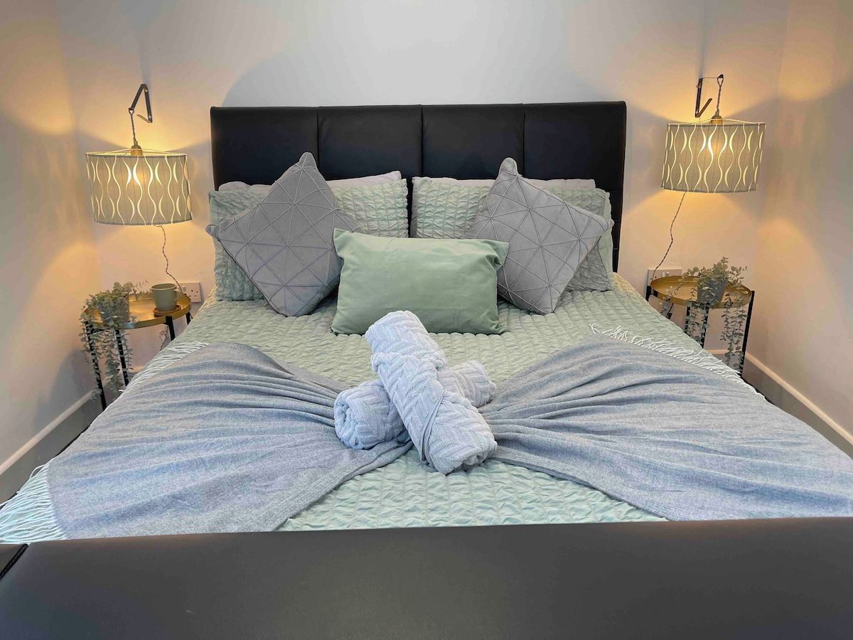 Ein ordentlich bezogenes Bett mit einem Kopfstück im geometrischen Muster, zwei Wandlampen und ein Paar gefaltete Handtücher auf dem Bett.