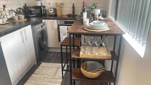 A compact kitchen with a built-in washing machine, a wooden table with wine glasses, and a window with blinds. - Cute+Cozy Guesthouse for 2 +secure offroad parking (Wolverhampton)