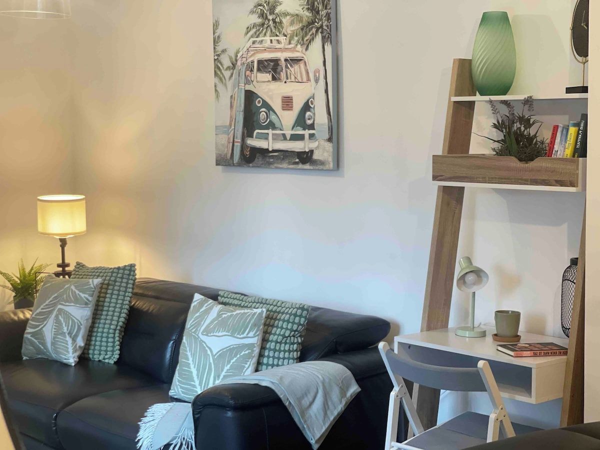 A living room with a black leather sofa, a painting of a vintage car, a lamp, a bookshelf, and a small table.