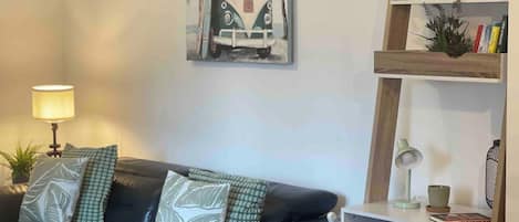 A living room with a black leather sofa, a painting of a vintage car, a lamp, a bookshelf, and a small table.