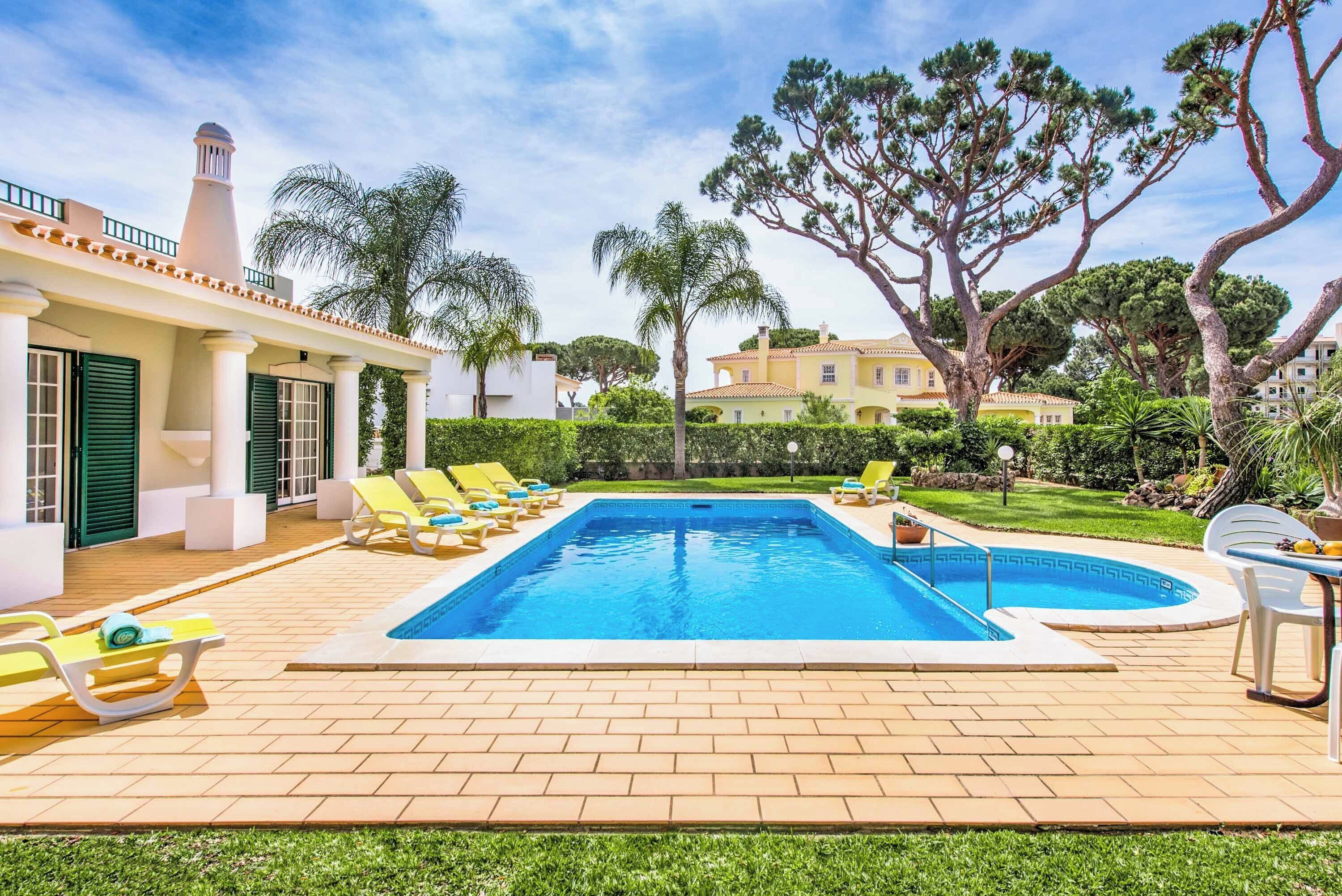 Une piscine avec des chaises longues, une terrasse carrelée et un grand arbre en arrière-plan.