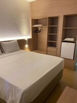 Quarto com uma cama grande, um guarda-roupa, um frigorífico pequeno e uma mesa de cabeceira com uma lâmpada.