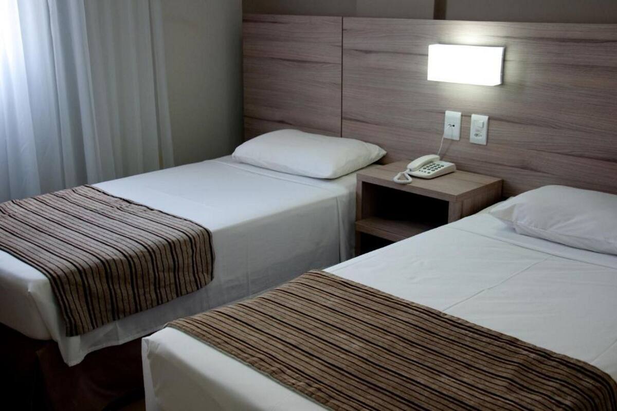 A hotel room with two beds, a bedside table with a phone, and a wall-mounted light.