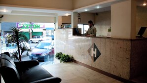 Zona de recepción de un hotel, con una recepcionista, un mostrador de recepción, un reloj en la pared y vistas a la calle.