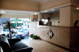 Uma área de recepção de hotel com recepcionista, balcão de atendimento e um relógio na parede, com vista para a rua.