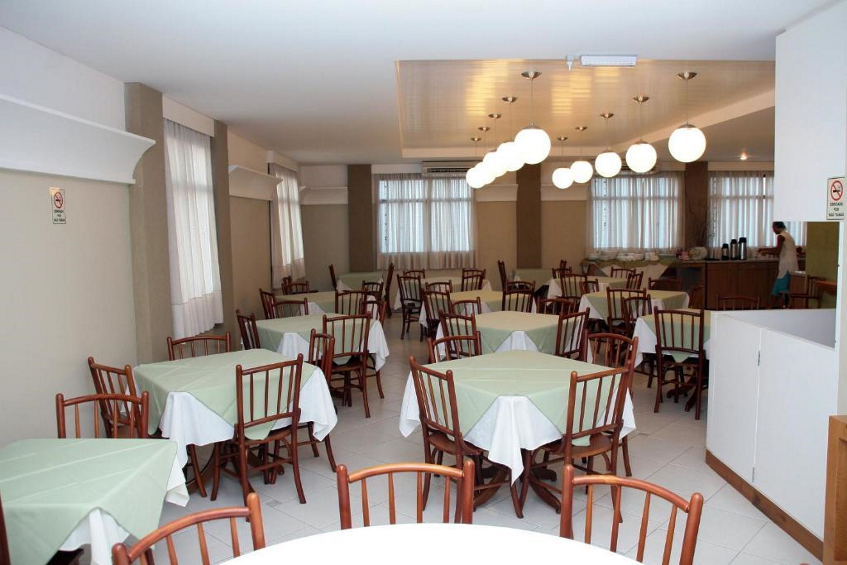 A dining area with multiple tables and chairs, a kitchen area, and a person working at a counter.