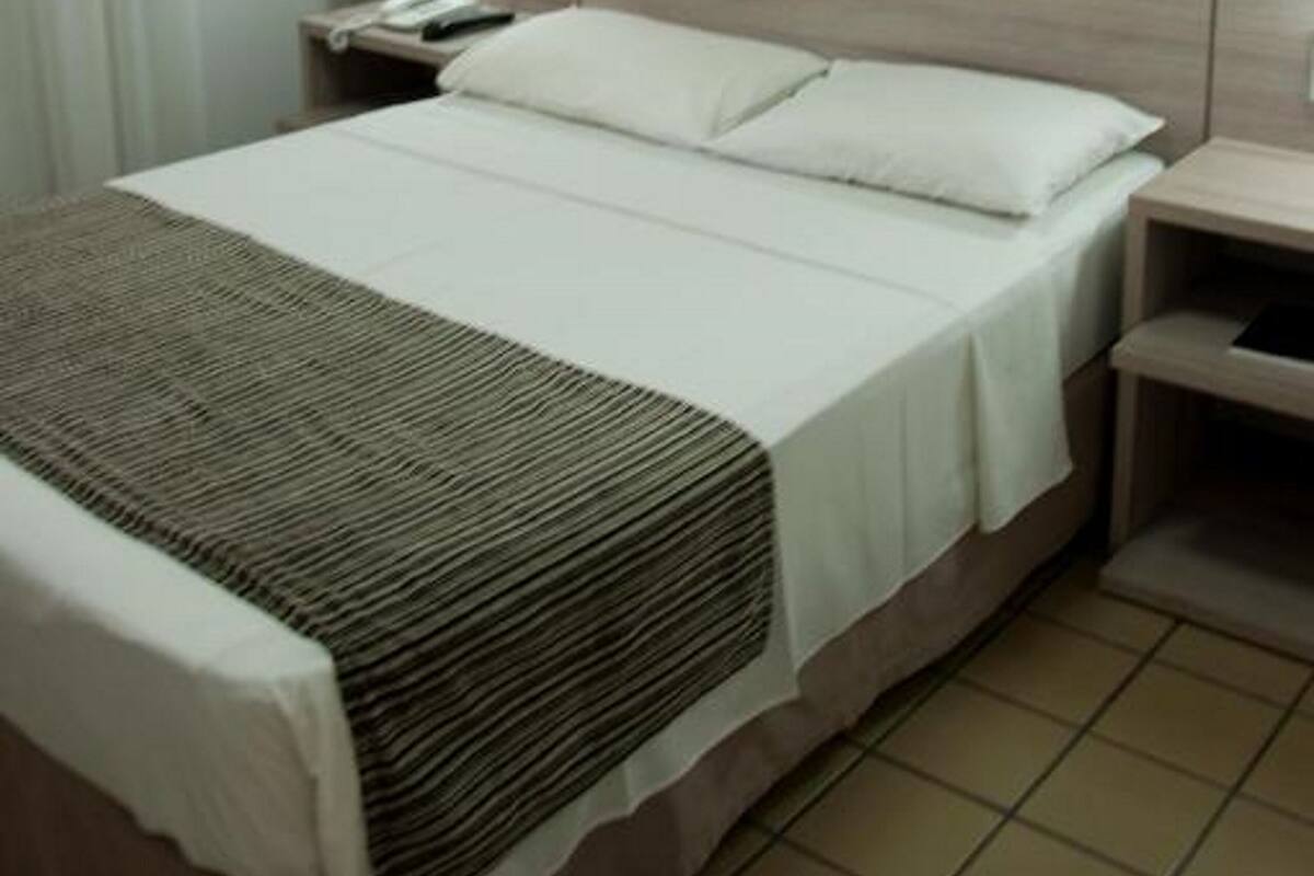 A hotel room with a bed, a nightstand, a wall-mounted heater, and a painting.