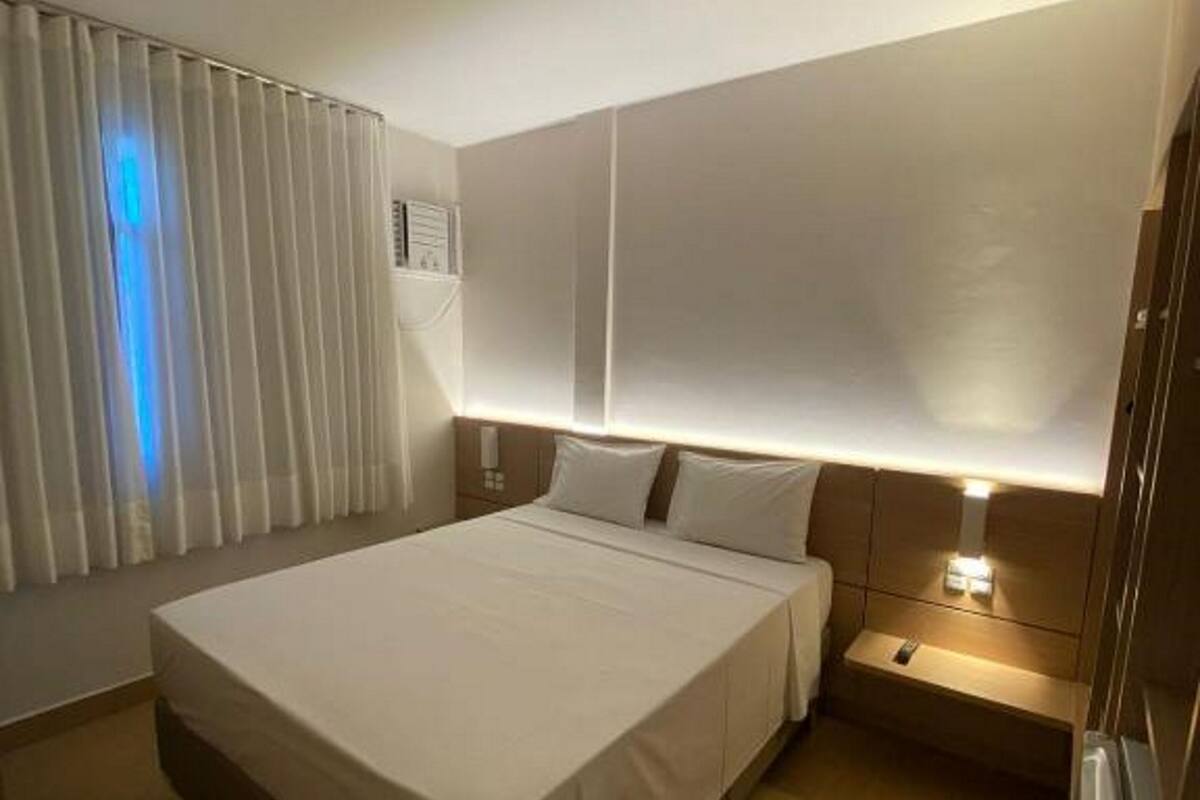 A hotel room with a large bed, a bedside table with a lamp, a window with blinds, and a ceiling light.