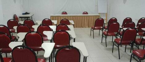 A conference room with rows of red chairs and white tables, a podium, and a whiteboard on the wall.