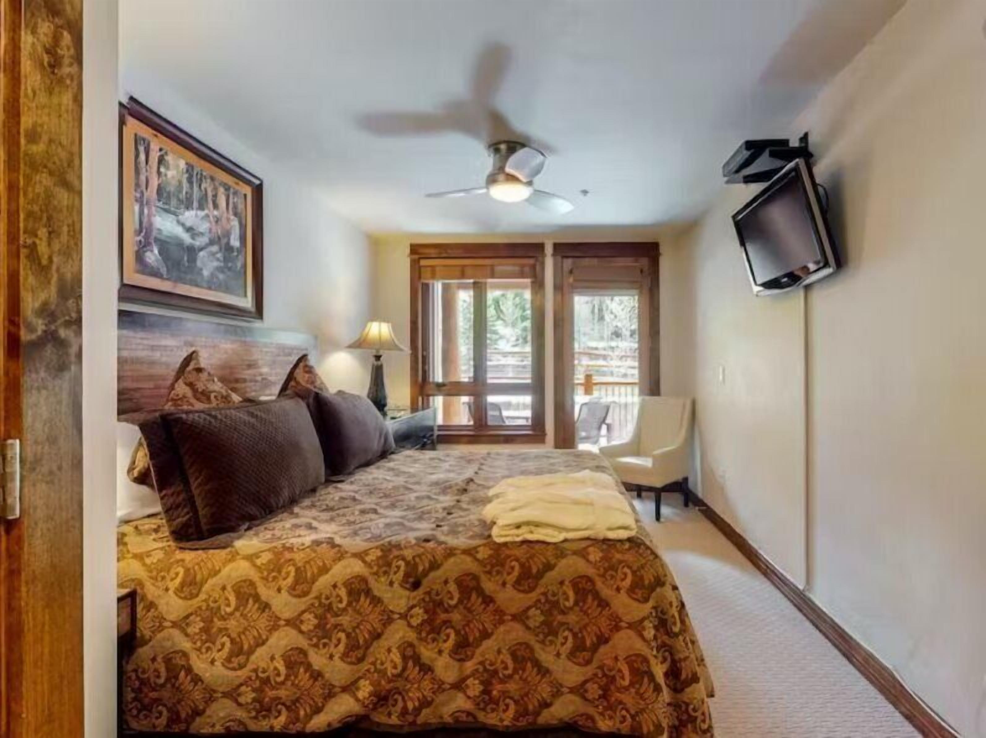 A bedroom with a bed, a ceiling fan, a TV, and a view of the outdoors.
