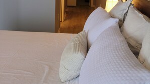 Une chambre d’hôtel avec un lit, des oreillers, une table de chevet et une lampe. Un couloir mène à une autre pièce.