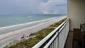 Une vue sur la plage depuis un balcon, avec un banc en bois, une jetée au loin et un ciel dégagé.