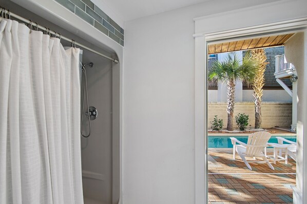 A bathroom with a shower, a white curtain, and a view of an outdoor pool area with chairs and palm trees.