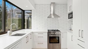 A modern kitchen with white cabinetry, a stainless steel range hood, and a large window providing a view of the outdoors. - Frankton Favourite - Queenstown Holiday Home (Queenstown)