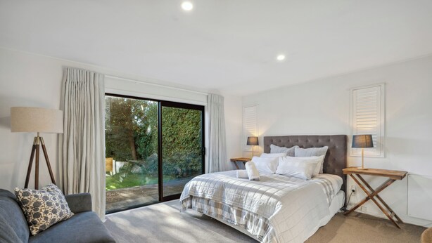 A bedroom with a bed, a sofa, a lamp, and a view of greenery through a sliding door. - Frankton Favourite - Queenstown Holiday Home (Queenstown)