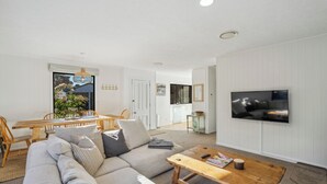 A modern living room with a sofa, coffee table, and TV. There is a dining area with a table and chairs. - Frankton Favourite - Queenstown Holiday Home (Queenstown)