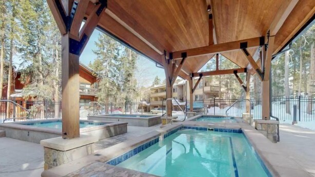 Zona de piscina exterior cubierta con varias piscinas, vigas de madera y vistas a edificios y árboles circundantes.