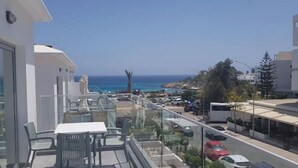 Un balcone con tavolo e sedie, affaccia su una zona costiera con edifici e auto parcheggiate.