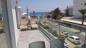 Un balcón con sofá y sillones, barandilla de vidrio y vistas a la calle y al mar.