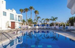 A swimming pool with lounge chairs and palm trees, surrounded by a white building with large windows. - Protaras Plaza Double Sea and Pool View-124 (Protaras)