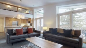 A modern living room with two sofas and a coffee table, a kitchen in the background, and large windows offering a view of trees.