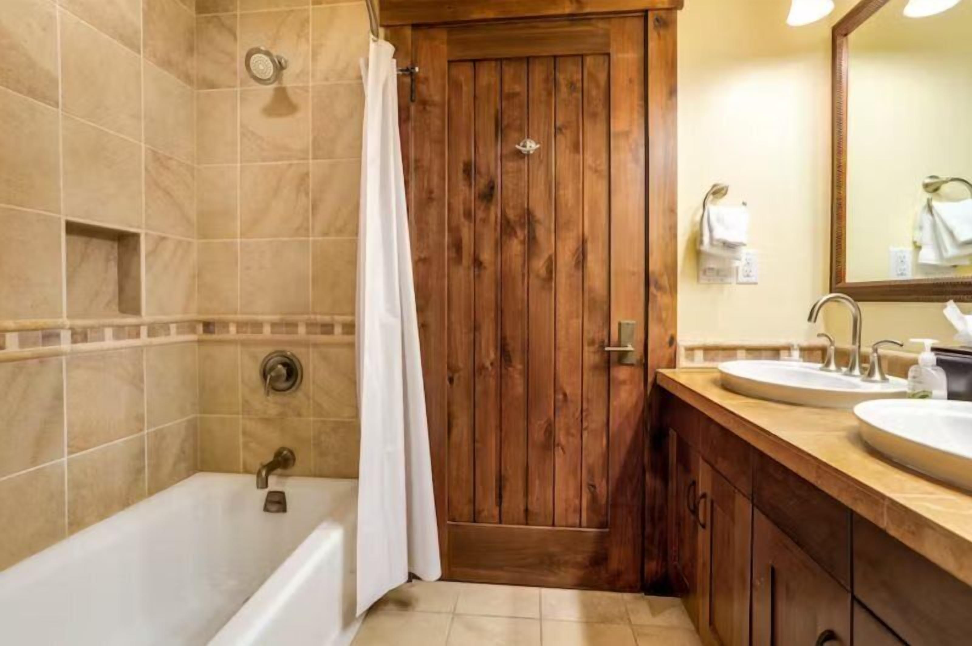 A bathroom with a wooden door, a bathtub, and a vanity with two sinks.