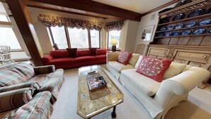 Una sala de estar acogedora con un sofá rojo, uno blanco y otro estampado. Hay una mesa de centro con revistas y una lámpara. La habitación tiene vigas de madera en el techo y una ventana con cortinas.