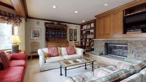 Una sala de estar acogedora con chimenea, un sofá, un sillón y una mesa de centro. Se observa una estantería con objetos decorativos y un televisor montado en la pared.