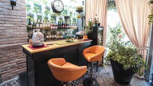 Zona bar con parete in mattoni, un orologio e diverse bottiglie. Sono presenti due sgabelli da bar arancioni e una grande pianta in vaso.