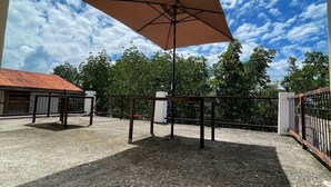 Un espace extérieur aménagé en terrasse, doté d’un grand parasol, d’un sol en gravier et offrant une vue sur des arbres et un bâtiment avec un toit rouge.
