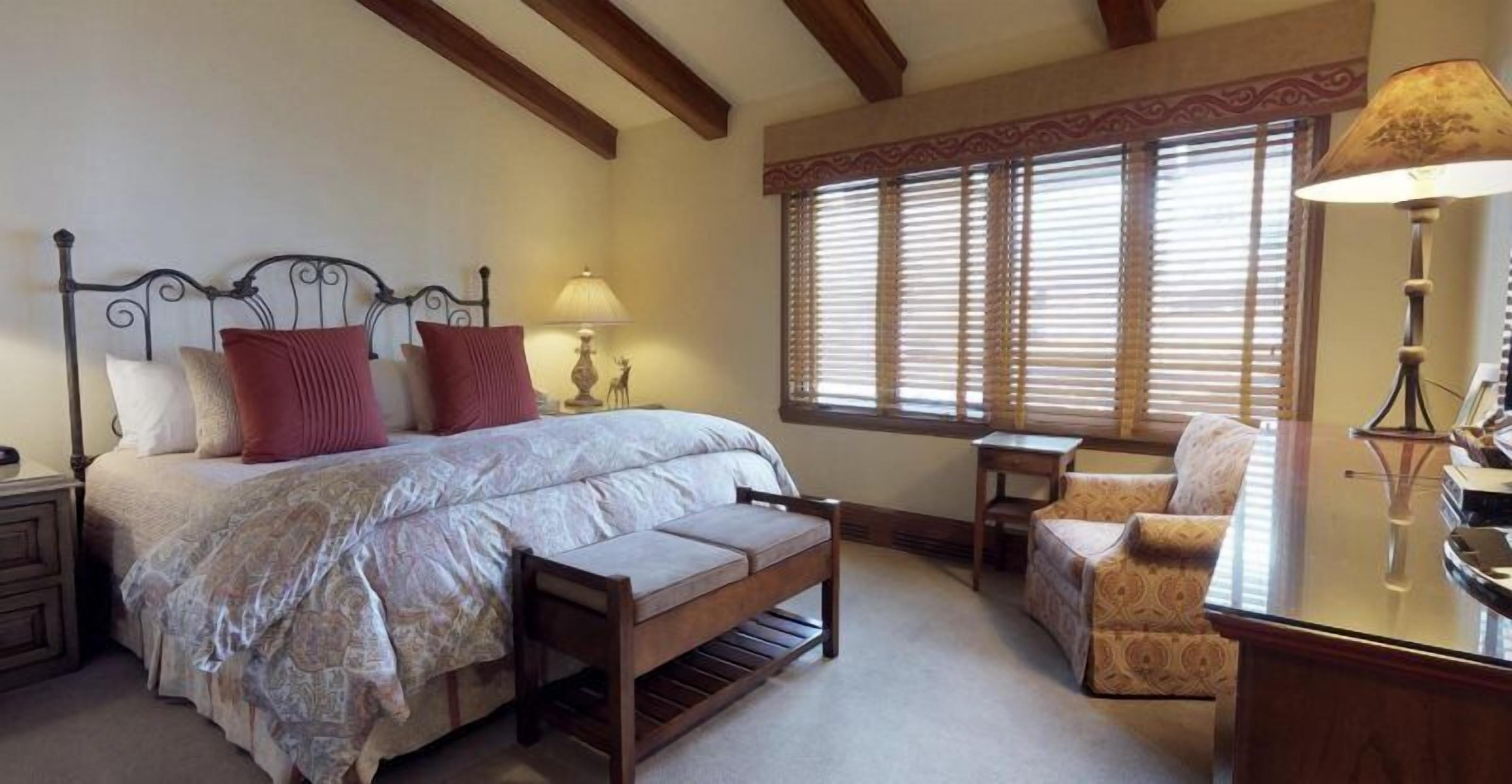 A bedroom with a bed, bench, chair, bedside table, lamp, and window with blinds.