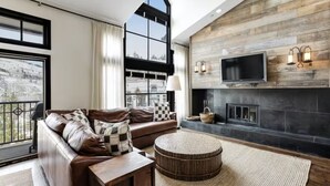 A modern living room with a leather sofa, a round coffee table, a fireplace, and a large TV mounted on a wooden wall. - 5 Bedroom Ski-in/ski-out Luxury Townhome in The Meadows Townhomes (Avon)