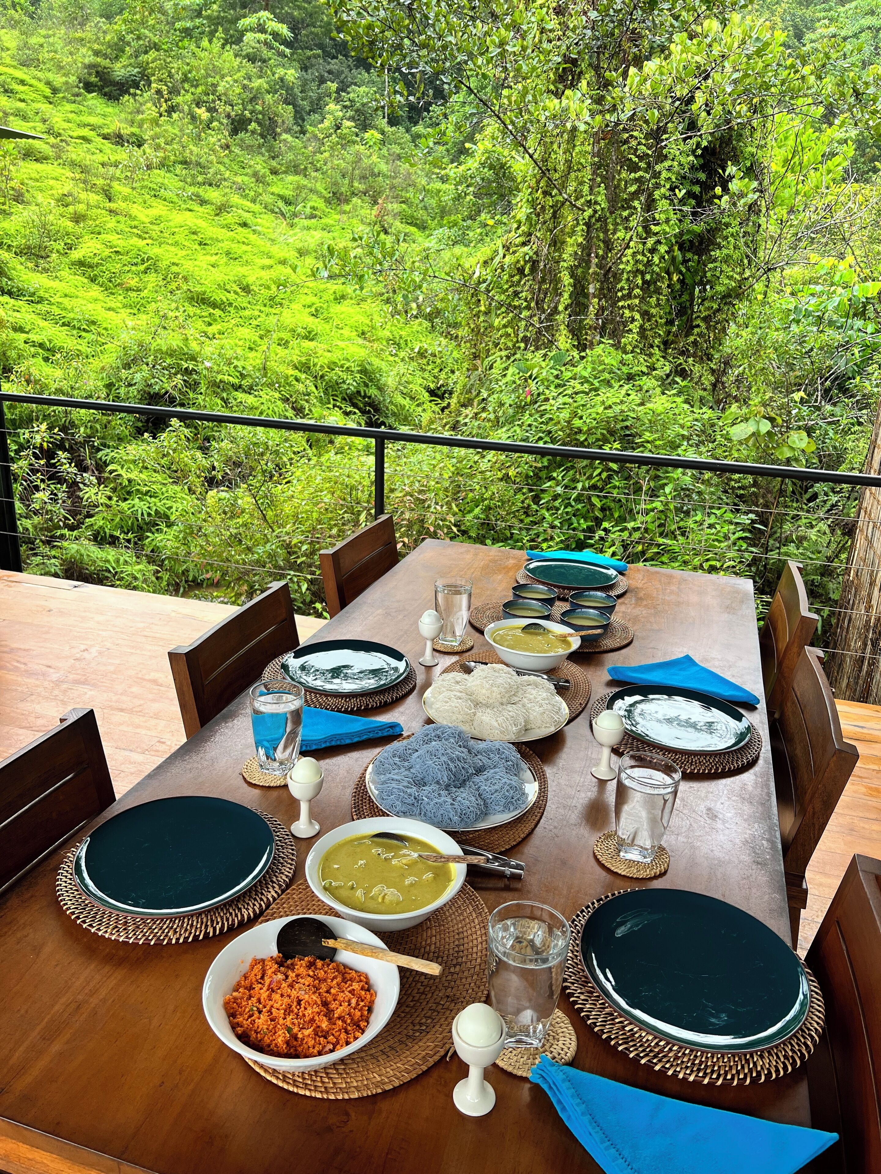 Une table à manger dressée avec différents plats et verres, entourée de chaises, et offrant une vue sur un paysage verdoyant et luxuriant.