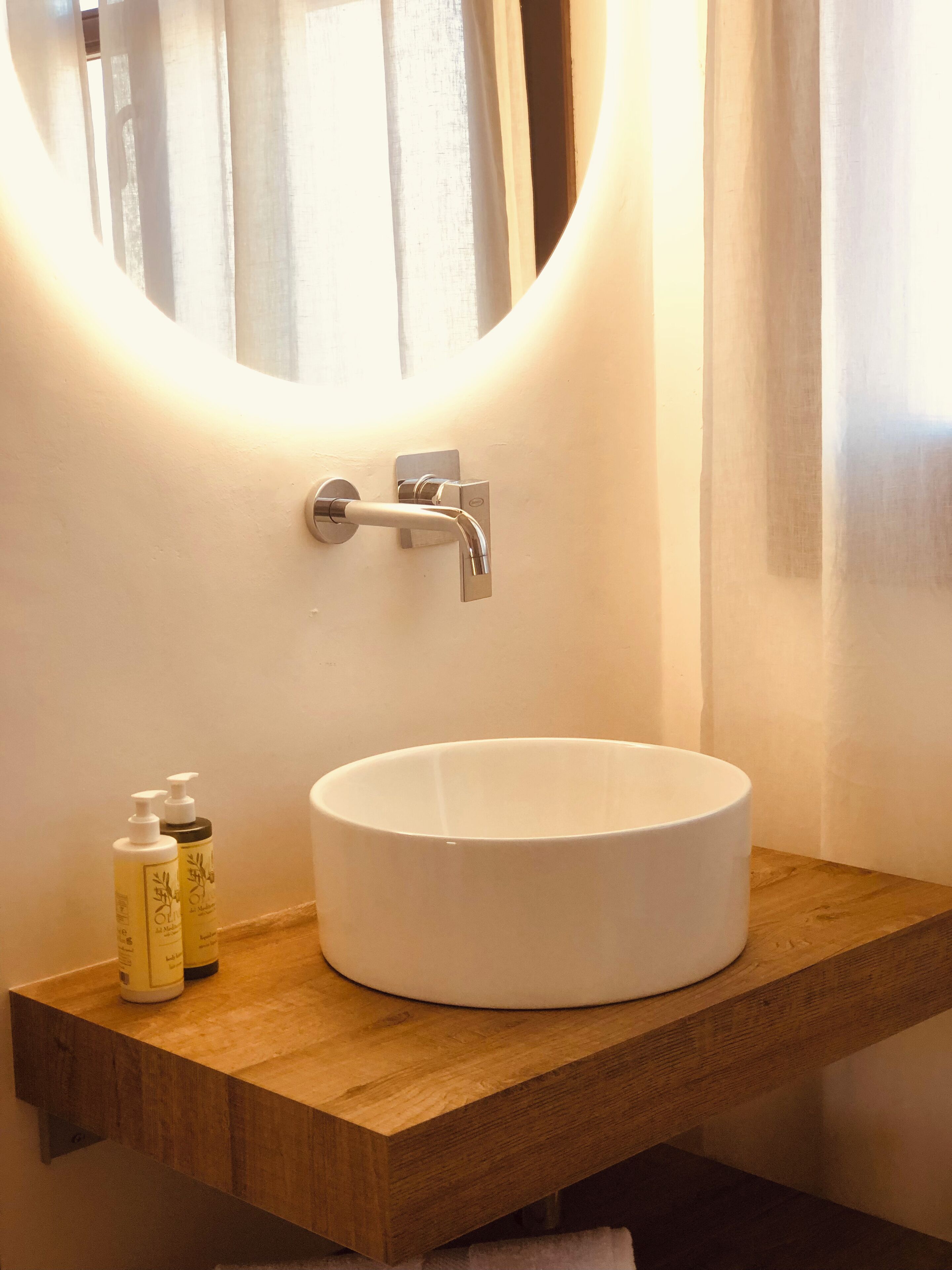 A bathroom with a round white sink, a wooden vanity, a mirror with a light, and two bottles of lotion.