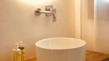 Un baño con un lavabo blanco redondo, un mueble de tocador de madera, un espejo con luz y dos botellas de loción.