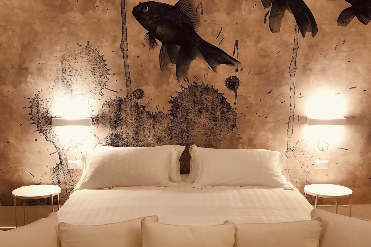 Cama com roupa de cama branca, duas mesas de cabeceira e um mural de peixes na parede.