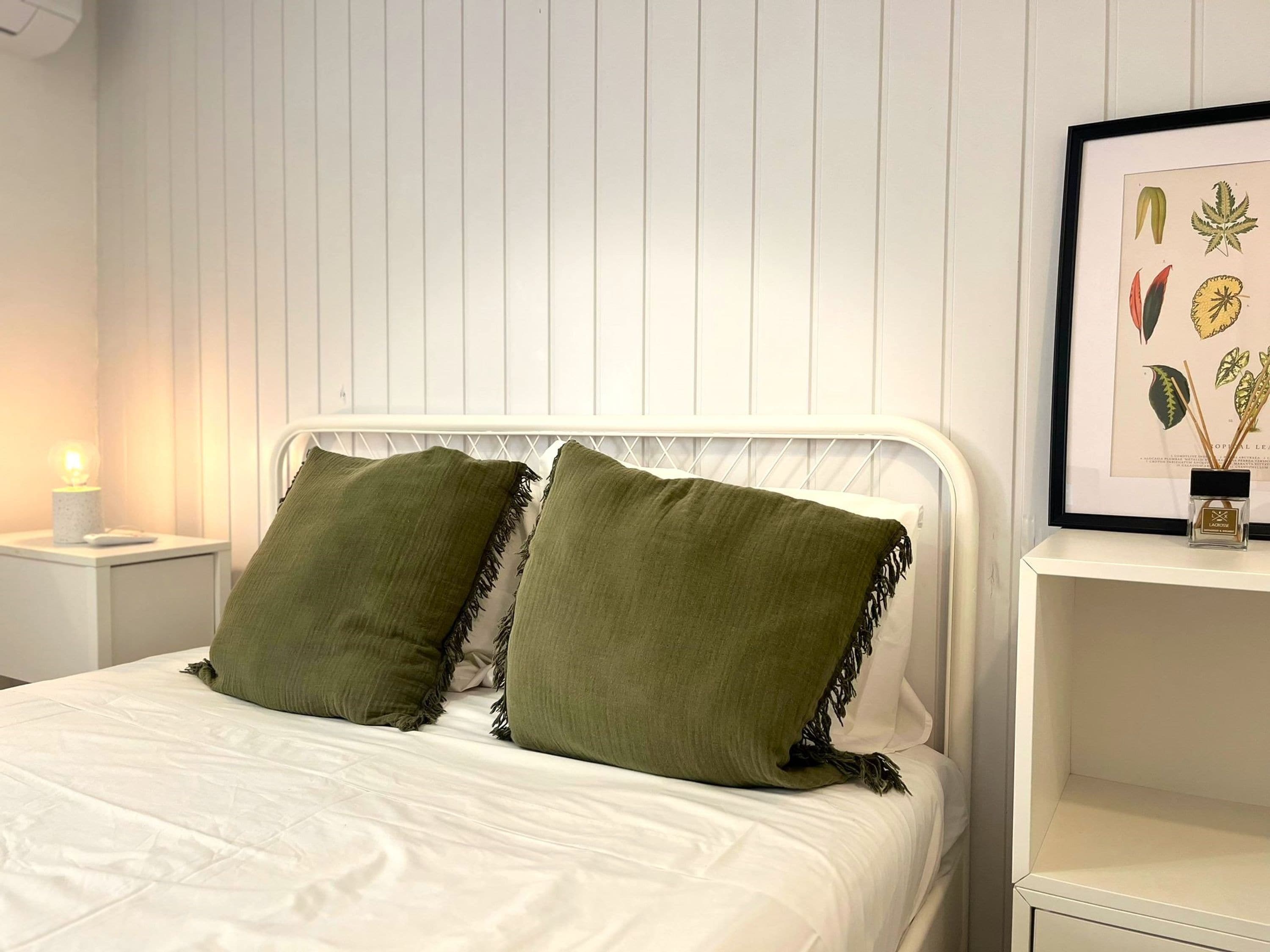 A neatly made bed with two green pillows, a white headboard, and a framed botanical illustration on the wall.