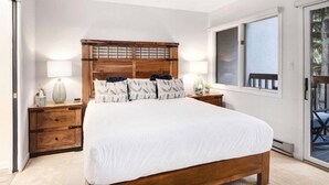 A bedroom with a wooden bed, white bedding, two bedside lamps, and a view of the outdoors through a sliding glass door.