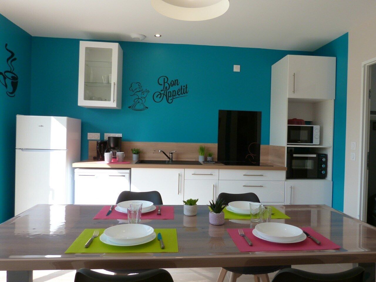 Une cuisine moderne aux murs de couleur turquoise, aux armoires blanches et dotée d’un coin repas aménagé pour quatre personnes.