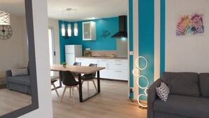 A modern kitchen with blue walls, white cabinets, and a black range hood. There is a dining area with a wooden table and chairs, and a gray sofa in the living room. - Domaine des Hameaux du Lac, 2 Chambres, 6 Pers max (SAINT PARDOUX LE LAC)