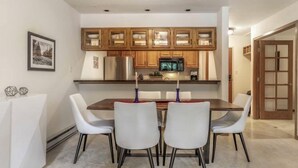 A dining area with a wooden table and white chairs, a kitchen with wooden cabinets and a refrigerator, and a door leading to another room. - Townsend Place 2 Bedroom Ski-in, Ski-out Condo in Beaver Creek Village (Avon)