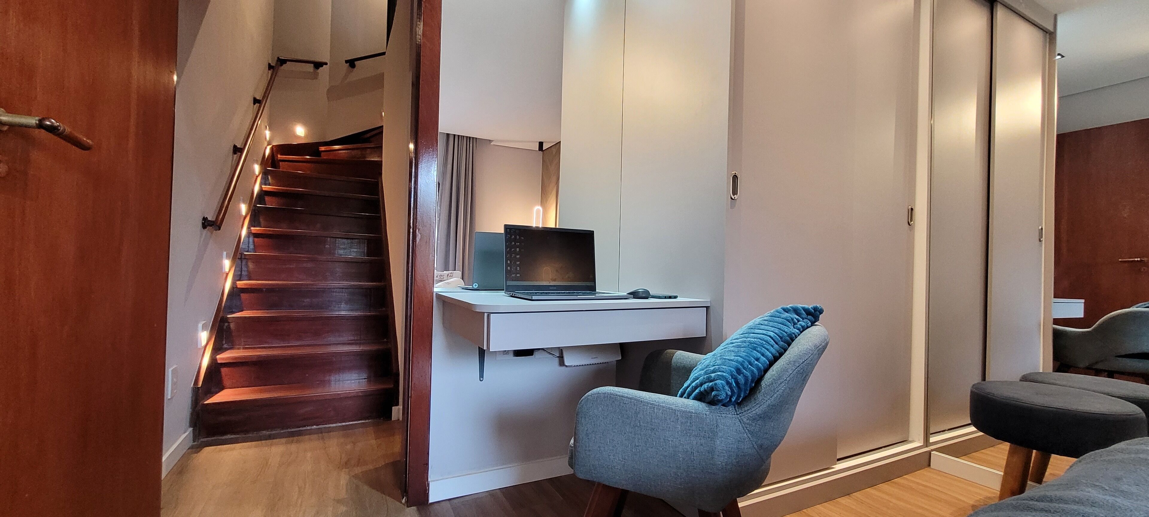 Um interior moderno com escada de madeira, uma escrivaninha com um laptop, uma poltrona cinza e um guarda-roupa.
