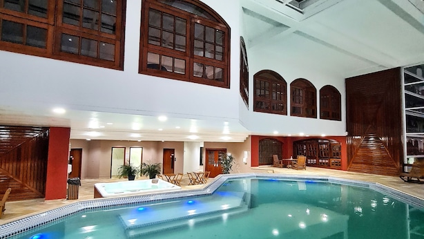 An indoor swimming pool with a glass ceiling, wooden windows, and a seating area with chairs and a hot tub. - Dúplex Luxo com Piscina Aquecida, Lareira e Wifi (Campos do Jordão)
