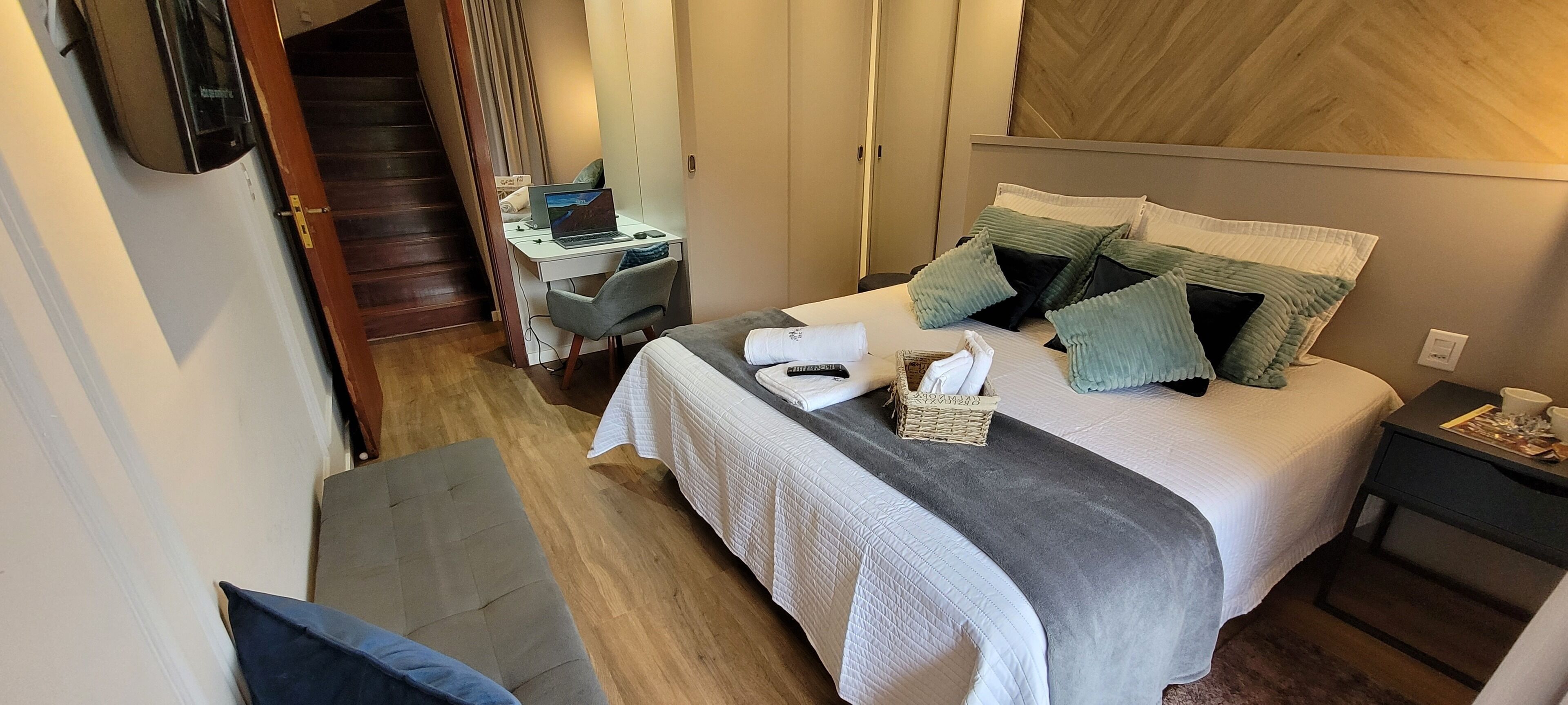 A bedroom with a bed, a desk with a laptop, a chair, a bedside table, and a staircase.