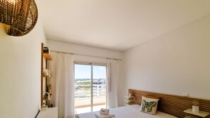 A hotel room with a large bed, a wooden headboard, a window with curtains, a view of the city, and a small shelf with toiletries.